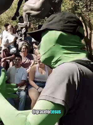 A person wearing a green face covering and a black hat is standing in front of a group of people. The person's arm is outstretched, seemingly gesturing at the group. The background includes greenery and a statue. The overall color scheme is natural, with the green of the face covering standing out.