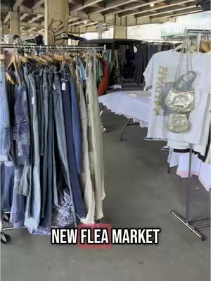 The image shows a busy flea market with racks of clothing. The scene is indoors under an industrial looking ceiling. On the left of the picture, racks of jeans and other clothes are visible. In the foreground, "NEW FLEA MARKET" is written in a black box. There are also racks with t-shirts on the right.