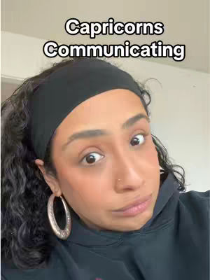 A woman with dark, curly hair, wearing a black hoodie and headband, and large hoop earrings, looks at the camera with a slight smirk. The background is a nondescript, light-colored wall. Overlayed text at the top reads, "Capricorns Communicating."