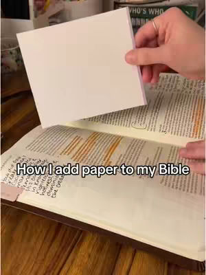 The cover image shows an open Bible on a wooden surface. A hand is holding a blank white piece of paper, about to insert it into the Bible. The image is well-lit, with a combination of warm and neutral tones. Text overlayed on the bottom of the image says, "How I add paper to my Bible".