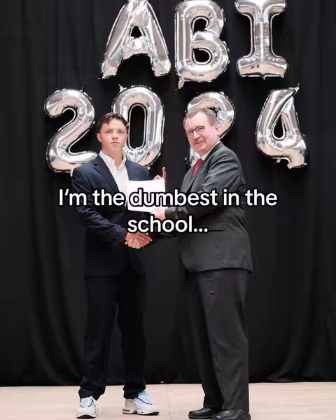 A young man in a suit is standing next to another man in a suit, both holding a certificate. Behind them are silver metallic balloons spelling "ABI 2024" against a black curtain.