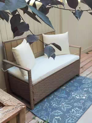 A patio scene features a wicker bench with white cushions and pillows set on a blue patterned rug. Overhead, leafy branches with dark leaves partially frame the scene, giving it a natural and cozy vibe.