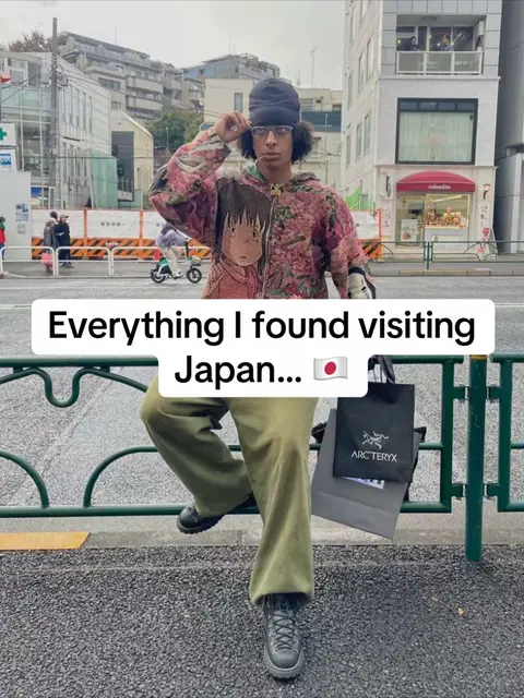 A person wearing a pink floral jacket, green pants, and a black beanie. They are leaning against a green metal fence with a gray sidewalk and building in the background. Text overlay says, "Everything I found visiting Japan..." with a Japanese flag emoji.