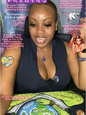 A woman is centered in a selfie-style video. She has light skin, dark eyebrows, and long, dark eyelashes, with full makeup. Her expression is open-mouthed as if speaking directly to the camera. She is wearing a black top with a plunging neckline. Behind her, a colorful background includes astrological symbols, text overlays, and small graphics. To the side, there's a small graphic of a cartoon girl, the colors are purplish. A microphone is clipped to her top, and a blue pendant hangs around her neck. The bottom of the frame is decorated with a green, decorated scarf.