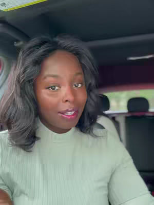 A medium shot shows a Black woman sitting in a car, looking directly at the camera and speaking. She has dark, wavy hair. She is wearing a light-colored top. The background shows the interior of the car and a slightly blurred view of the outside. The lighting is natural.
