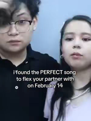 The cover image shows a young couple, a man and a woman, in a medium shot taken indoors. The man on the left is wearing glasses and a black polo shirt, while the woman is on the right with a white shirt and is wearing a pearl necklace. Both look directly at the camera. The background is a plain, light blue wall.