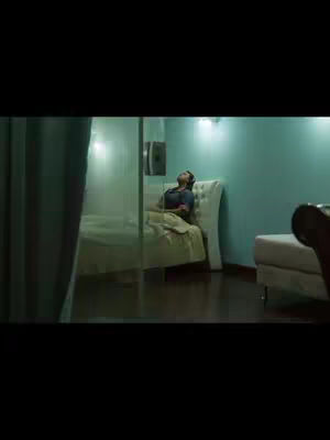 A room interior shot with a person lying on a bed. The room is dimly lit, with a turquoise wall and a white bed highlighted. There is a glass wall on the left side. The person appears to be lying on their back on the bed with their eyes closed.