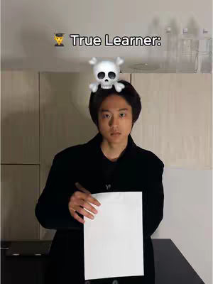 A young man stands centered in the frame, holding a blank white paper in front of him. He's dressed in a black jacket, and a small white skull with crossbones sits on top of his head, positioned as an overlay. Above him is a text overlay that reads 'True Learner: ' along with a graduation cap emoji. The background consists of neutral-colored walls and a dark surface, with slight variations in the lighting across the scene.