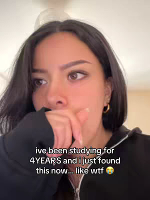 A close-up shot of a woman with dark short hair, slightly covering her forehead. She's wearing a black zip-up hoodie and gold hoop earrings. Her right hand is raised, with her fingers near her lips as if she's deep in thought or surprised. The background is soft, with blurred neutral tones.