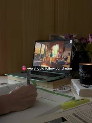 A laptop displays a scene of a room, with a person in the background. In the foreground, a hand holds a pen above an open book. Other items like a pen, highlighter, and a candle are also there, with a study setup.