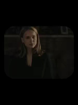 A medium shot shows a young woman standing in a dark, dimly lit space. She has light skin and shoulder-length blonde hair. She wears a dark top. Her gaze is directed towards the camera with a somewhat serious expression. The background is out of focus, providing a sense of mystery or introspection.