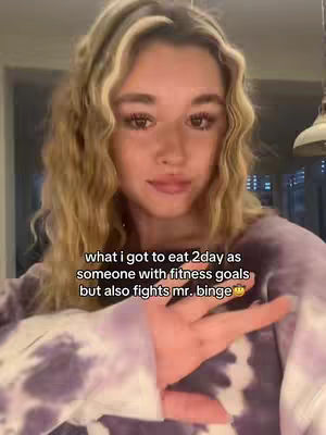 A young woman with long, wavy blonde hair is centered in the frame. She wears a purple tie-dye sweatshirt and is positioned in front of a neutral background. The lighting seems soft and natural, illuminating her face and upper body. The image's composition is a medium-shot, focusing on the woman's expression and the overlay text that appears on the bottom. She is gesturing with her right hand, keeping the focus on the hook.