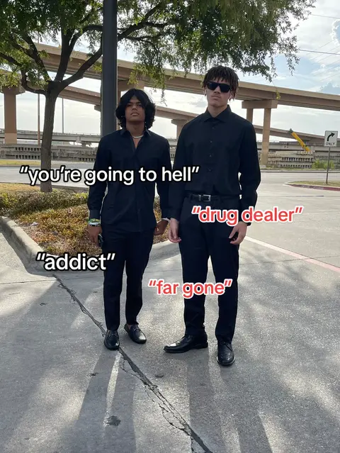 Two young men stand on a paved area, likely a sidewalk or parking lot, in front of a blurred background featuring buildings and a cloudy sky. Both are dressed in black button-down shirts and dark pants. The man on the left has long dark hair, while the man on the right wears sunglasses. Text overlays appear near them, like dialogue labels, expressing negative self-perceptions.