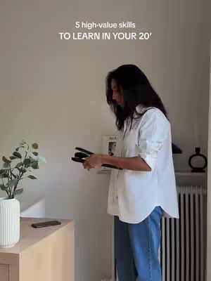A woman in a white button-up shirt and blue jeans stands indoors holding a hair styling tool near a wall. The upper section of the image has text overlayed, and the surroundings include furniture and decor.