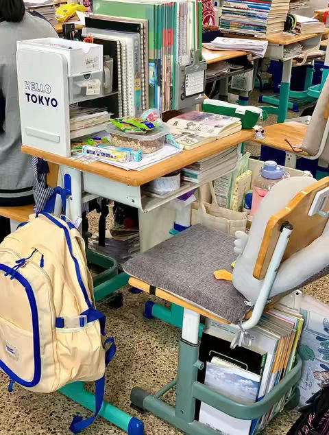 A cluttered desk in a classroom setting, filled with books, notebooks, study materials, and a backpack.