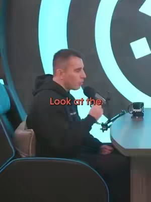 A man with short hair, wearing a black hoodie, is seated in a chair behind a desk with a microphone, looking to his right while speaking. The background features a dark wall with a blue circular pattern. The text overlay, "Look at the", is in red.