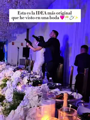 A photo taken at a wedding. The foreground features a table decorated with white flowers, candles, and glassware. In the background, a bride and groom are aiming toy guns at the camera, with a party backdrop.