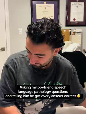 A man is seated and looking down. He has dark curly hair and is wearing a dark gray t-shirt. The text overlay states he is answering speech pathology questions.