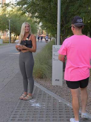 A woman stands on a paved path, holding a beverage and looking towards a man standing in the background. She's wearing a black top and grey leggings. The man is wearing a pink shirt and black shorts. The scene takes place outdoors, with trees and a building visible in the background.