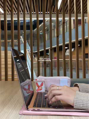 A person's hands typing on a pink laptop, with a pink laptop case and a pencil case in the background. The setting seems to be a modern library or co-working space, with wooden slatted ceiling and walls of glass.