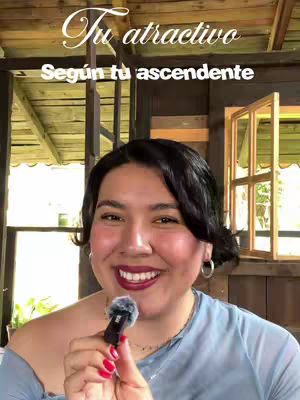 A woman with black hair smiles and holds a microphone; text overlays declare: 'Tu atractivo, Según tu ascendente' (Your attractiveness, According to your sign). A background of wooden structure adds a rustic vibe.