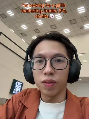 A selfie-style image featuring a person in glasses wearing headphones. The background is an indoor ceiling with lights, and there's a sign with text in the background.