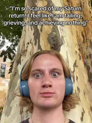 A selfie-style shot of a young man with surprised eyes and a slightly open mouth. He wears blue over-ear headphones and is positioned in front of a large tree trunk with textured bark.
