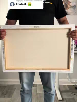 A man in a black shirt and light blue jeans holds a large, blank, white-framed canvas on a stand, the back side facing towards the camera. His arms and torso fill most of the frame, with a partial view of a comment overlayed at the top left corner. Soft light suggests an indoor setting.