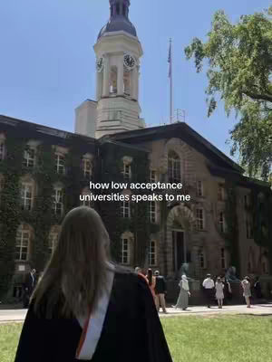 A woman in a graduation gown stands with her back to the camera, facing a grand stone building with a clock tower. The sky is clear blue. The text overlay reads how low acceptance universities speaks to me.