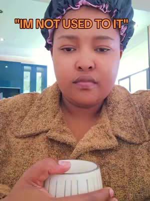 A woman in a brown jacket and purple head covering holds a white cup. The text overlay reads "I'M NOT USED TO IT" in orange letters.