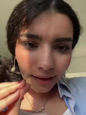 A close-up shot of a woman looking down slightly with a neutral expression, her face illuminated with soft lighting. She has dark hair and is wearing a light blue shirt, with a necklace visible. The background is softly blurred.