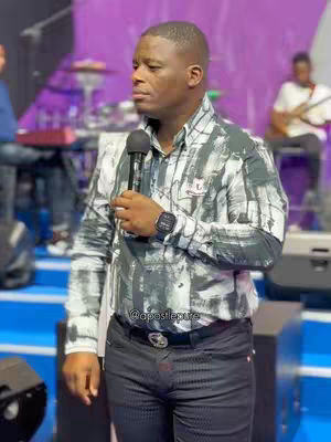A man in a black-and-white patterned shirt is holding a microphone and standing on a stage. The background is blurred, but there is a person playing the guitar and a drum kit. The color scheme is purple and blue.