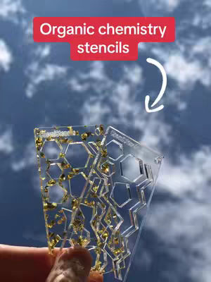 A hand holding two clear stencils with hexagonal cutouts in front of a cloudy blue sky. A red rectangle with white text that reads "Organic chemistry stencils" is at the top, with a curved white arrow pointing towards the stencils.