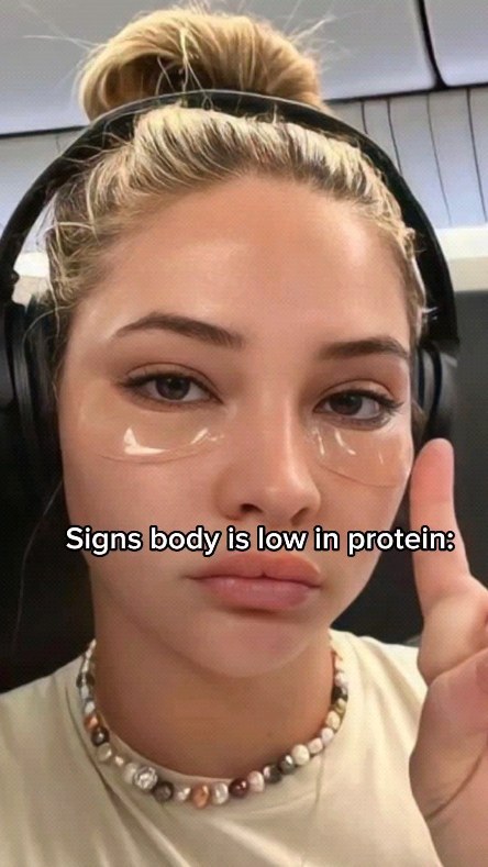 A selfie-style image featuring a young woman with blonde hair in a bun and headphones around her neck. She has under-eye patches, suggesting a skincare routine. The background is blurred, and she has a necklace made of various beads and stones. The text overlay reads, 'Signs body is low in protein:'.