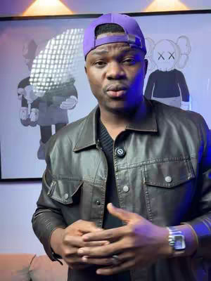 A man in a purple cap and dark jacket speaks to the camera. Behind him, there is artwork displayed on the wall.