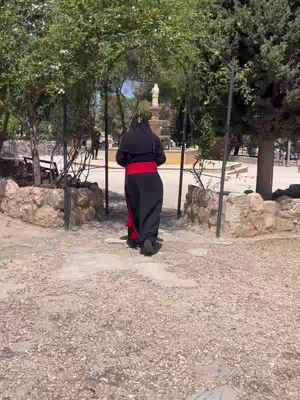 A person in black robes with a red sash is walking towards a fountain in a public park. They are seen from the back, and the scene is framed by trees and a stone archway.