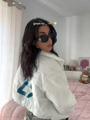 The cover image features a woman in a room, posing. She is wearing sunglasses and a white jacket. Her hair is dark and styled. The background includes a bed with a pink comforter and a window.