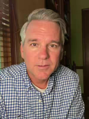 A middle-aged man with gray hair and a blue checkered shirt is centered in the frame, with a neutral expression. Behind him, a domestic setting includes window blinds on the left and a glimpse of a bookcase on the right.