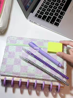 The image showcases a well-organized workspace featuring a laptop, a notebook, pens, and sticky notes. The notebook has a checkered pattern, while the pen and notebook share a color scheme of lavender and purple. The layout suggests a focus on productivity and organization