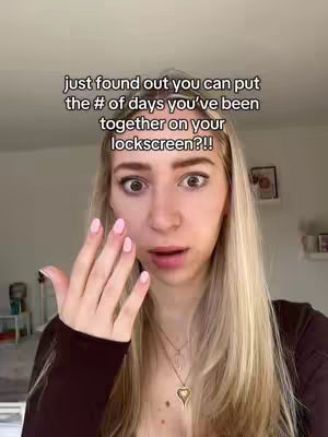 A woman with long blonde hair and fair skin looks surprised, with wide eyes and a hand raised towards her face. Text overlay reads 'just found out you can put the # of days you've been together on your lockscreen??!!'. Background is a blurred interior with hints of a living space.