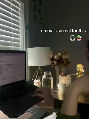 A well-lit room with a laptop on a desk. The laptop screen displays text, and a person is using the trackpad with the right hand. There is a window with blinds, and a table lamp is on the desk near a vase of flowers and other decorative items. The added text says: emma's so real for this. The image conveys a sense of studying.