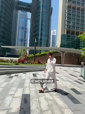 A man in a white outfit stands on a tiled area of a modern city, talking into his phone. Tall, contemporary buildings form the background under a clear blue sky. The image is well-lit, showing a sunny day with a focus on urban surroundings.