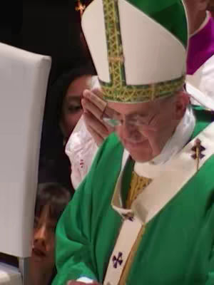 The image shows Pope Francis wearing a green vestment and white mitre, with his hands together in prayer. Other people are visible in the background. The composition is a medium shot, focused on the Pope's face. The lighting is soft and natural.