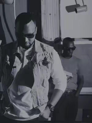 Two men wearing sunglasses stand closely together. The foreground man, wearing a denim jacket and white shirt, has his hand in his pocket and is looking down. The second man is behind, slightly out of focus wearing a light colored t-shirt. The background is an indistinct mix of shapes and patterns, mostly in shades of gray.