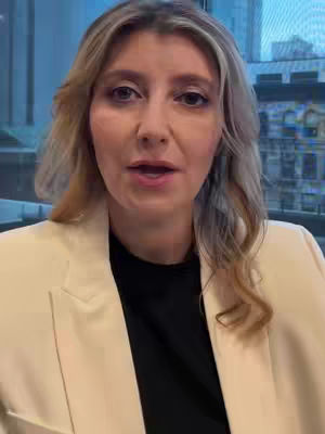 A woman with blonde hair and a cream-colored blazer is shown from the chest up, facing the camera and speaking. Behind her is a blurred view of a building with large windows. The lighting appears to be natural and soft, with the focus clearly on the woman.