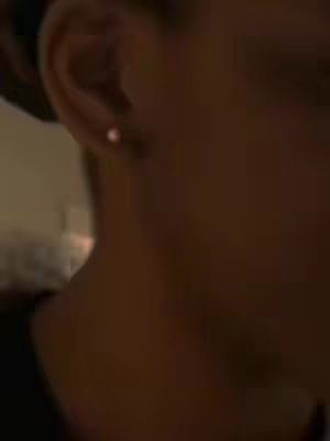 A close-up shot of a person's ear with a small stud earring. The skin tone is medium-dark. The background is out of focus and appears to be a dimly lit room.