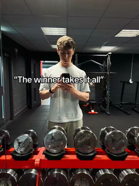 A man in a gym holding a phone up taking a mirror selfie in front of a set of free weights.