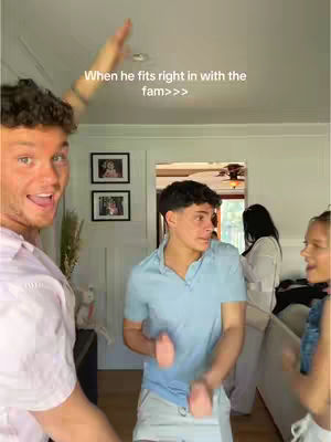 The cover image captures a lively scene with three individuals in a well-lit indoor setting. A man on the left is smiling widely, while gesturing with his right arm towards the upper middle of the frame. Another man in the center is looking slightly off to the side with a neutral expression. A woman is partially visible on the right side, also smiling. The background reveals a glimpse of a door, framed pictures, and additional figures, suggesting a casual, familial atmosphere. The text overlay reads, "When he fits right in with the fam>>>", adding context to the interpersonal dynamic.