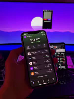 A hand holding a smartphone displaying a cryptocurrency trading app interface. The background shows a monitor with a purple gradient and what appears to be a desk setup with another monitor. The phone screen showcases account balance and various cryptocurrency icons.