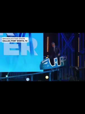 A man in a suit rides a small amusement park ride. Behind him is a large screen with a blue hue and the text "ER". Below the screen, on the ride's path are lights for a show.
